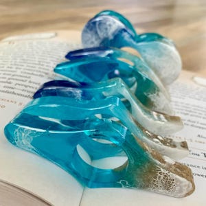 May include: A set of four blue and white resin bookmark rings. The rings are shaped like waves and have a layered effect, with white at the top and blue at the bottom. The rings are arranged in a row, with the largest ring at the back and the smallest ring at the front.