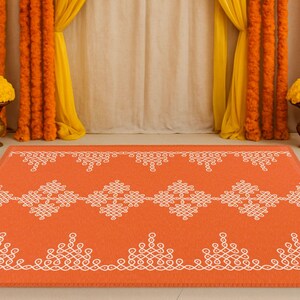 May include: An orange and white patterned rug with a traditional Indian design. The rug is placed in front of a backdrop of yellow and orange flower garlands and white curtains.