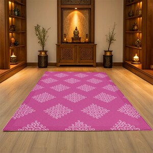 May include: A pink area rug with a white geometric pattern. The rug has a diamond shape with a white design inside each diamond. The rug is on a wooden floor in a room with a wooden cabinet and shelves.