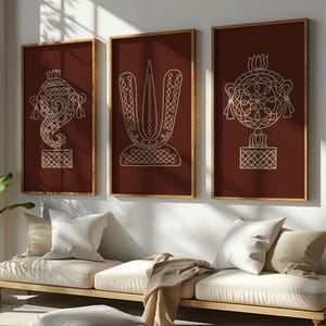 May include: Three framed wall art pieces with intricate white line drawings on a deep red background. The art features geometric and symbolic designs, including a stylized elephant, a vertical emblem, and a circular pattern. The frames are a warm, natural wood tone.