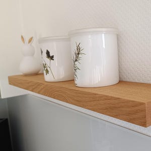K&ouml;nnte beinhalten: Ein hellbraunes Holzregal zeigt zwei wei&szlig;e Keramikgl&auml;ser mit botanischen Designs und Deckeln sowie eine wei&szlig;e Hasenfigur. Das Regal ist auf einer hellgrauen Oberfl&auml;che vor einer strukturierten wei&szlig;en Wand montiert.