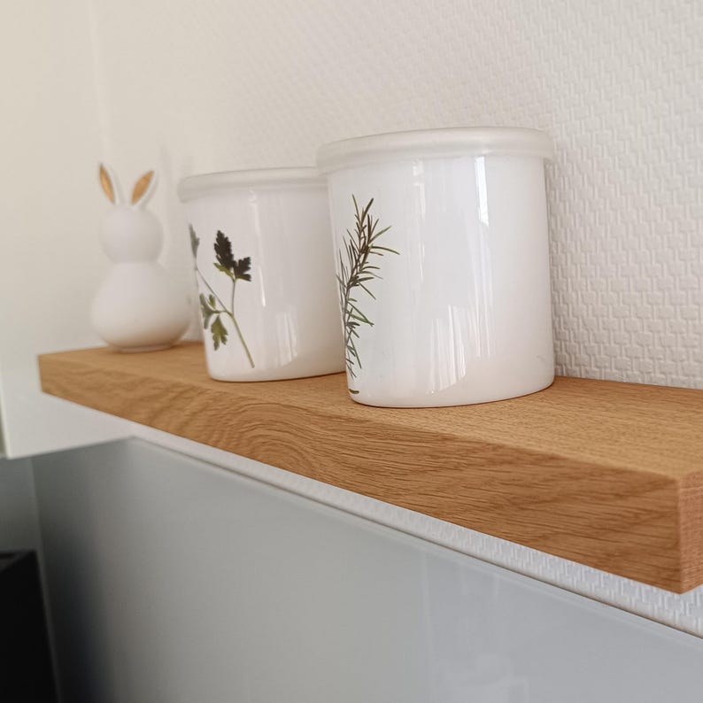 K&ouml;nnte beinhalten: Ein hellbraunes Holzregal zeigt zwei wei&szlig;e Keramikgl&auml;ser mit botanischen Designs und Deckeln sowie eine wei&szlig;e Hasenfigur. Das Regal ist auf einer hellgrauen Oberfl&auml;che vor einer strukturierten wei&szlig;en Wand montiert.