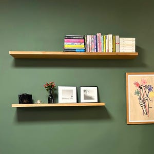 K&ouml;nnte beinhalten: Zwei schwebende Holzregale an einer gr&uuml;nen Wand. Das obere Regal enth&auml;lt eine Sammlung von B&uuml;chern mit farbigen Buchr&uuml;cken. Das untere Regal zeigt eine kleine schwarze Kamera, eine Vase mit roten Blumen, zwei gerahmte Fotos und eine Kerze.