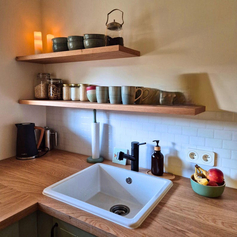 K&ouml;nnte beinhalten: K&uuml;chenszene mit Holzregalen, die Gl&auml;ser, Tassen und Schalen tragen. Ein wei&szlig;es Waschbecken steht auf einer Holz-Arbeitsplatte, zusammen mit einem schwarzen Wasserhahn und Seifenspender. Eine Obstschale und ein schwarzer Wasserkocher sind ebenfalls zu sehen.
