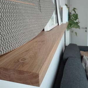 K&ouml;nnte beinhalten: Ein hellbraunes Holzregal mit nat&uuml;rlicher Holzmaserung. Ein dekorativer Gegenstand mit grauem und wei&szlig;em Muster liegt auf dem Regal. Das Regal ist an einer wei&szlig;en Wand befestigt, mit einem Blick auf ein graues Sofa im Hintergrund.