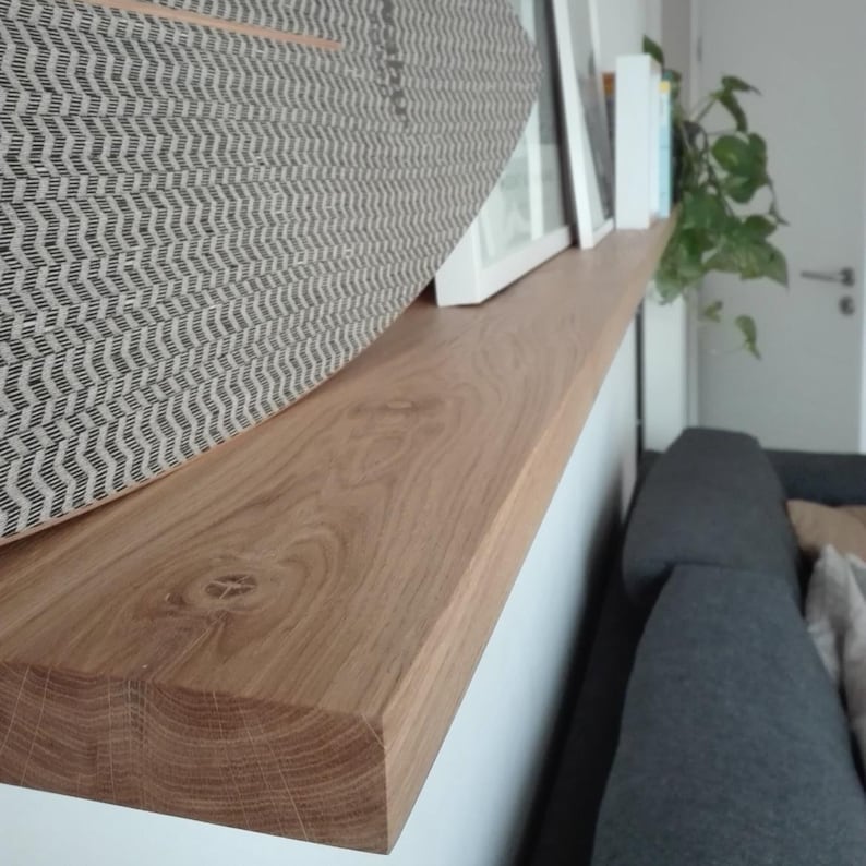 K&ouml;nnte beinhalten: Ein hellbraunes Holzregal mit nat&uuml;rlicher Holzmaserung. Ein dekorativer Gegenstand mit grauem und wei&szlig;em Muster liegt auf dem Regal. Das Regal ist an einer wei&szlig;en Wand befestigt, mit einem Blick auf ein graues Sofa im Hintergrund.