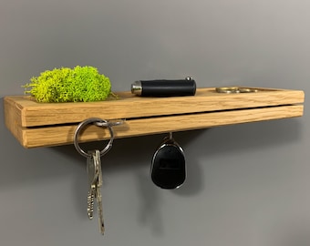 Könnte beinhalten: Ein hölzerner Schlüsselhalter mit einem kleinen Regal. Das Regal enthält ein schwarzes Messer, einen kleinen Haufen Münzen und einen Flecken grünes Moos. Schlüssel und ein Autoschlüsselanhänger hängen darunter. Das Holz hat eine natürliche Maserung.