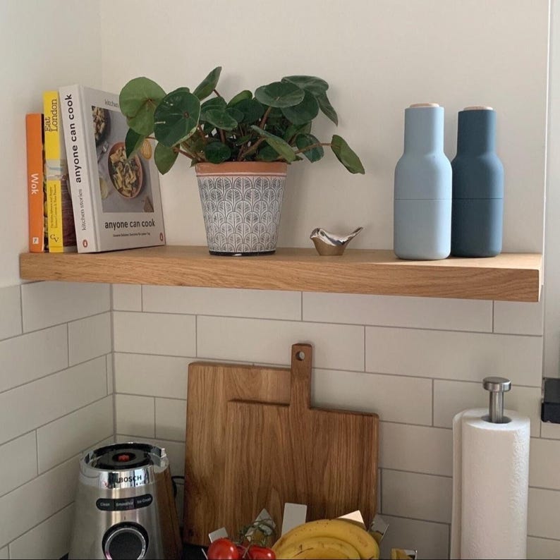 K&ouml;nnte beinhalten: Ein Holzregal zeigt eine Topfpflanze, Kochb&uuml;cher und zwei blaue M&uuml;hlen. Eine kleine Metallvogel-Figur sitzt neben der Pflanze. Darunter sind ein Holzschneidebrett und ein Mixer zu sehen, was auf eine K&uuml;chenumgebung hindeutet.