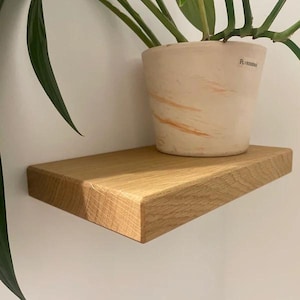 K&ouml;nnte beinhalten: Ein schwebendes Regal aus Naturholz mit abgeschr&auml;gter Kante, das einen beigefarbenen Keramik-Pflanzentopf mit orangefarbenen Pinselstrichen tr&auml;gt. Das Regal ist an einer wei&szlig;en Wand befestigt, gr&uuml;ne Bl&auml;tter sind sichtbar.