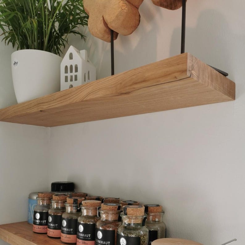 K&ouml;nnte beinhalten: Ein Holzregal mit nat&uuml;rlicher Oberfl&auml;che, an einer wei&szlig;en Wand befestigt. Das Regal enth&auml;lt eine Topfpflanze, eine kleine wei&szlig;e Hausfigur und mehrere Gl&auml;ser mit Korken. Die Gl&auml;ser enthalten Gew&uuml;rze.