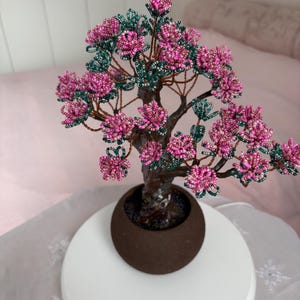 May include: A decorative bonsai tree sculpture crafted from wire and small beads. The tree features a textured brown trunk and branches adorned with clusters of pink and dark green beads, arranged in a round, dark brown ceramic pot base.