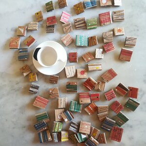 Può includere: Una vista dall'alto mostra una collezione di piccole opere d'arte rettangolari dipinte a mano, sparse attorno a una tazza e un piattino bianchi. Le opere presentano vari disegni astratti nei colori rosso, verde e blu, su un materiale simile al cartone.