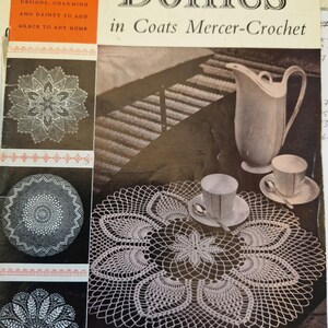 Puede incluir: Una portada de libro vintage con un encaje de ganchillo blanco sobre un fondo oscuro. El título del libro es "Doilies in Coats Mercer-Crochet" y el precio es de 15p. La portada del libro también presenta una jarra blanca y una taza de té blanca con su platillo.
