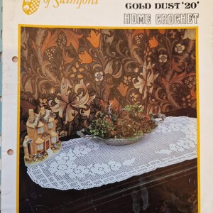 May include: A white crocheted table runner with a floral design. The runner is on a dark wood table with a small house figurine and a bowl of greenery. The image is from a vintage crochet pattern book titled "Twenty or Gold Dust '20' Home Crochet" by Twilleys of Stamford.
