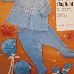 May include: A light blue knitted baby set, including a jacket, pants, two hats, and mittens. The jacket has button closures and the pants have an elastic waistband. The set is designed for a 16 to 20 inch chest. The Hayfield brand is visible.