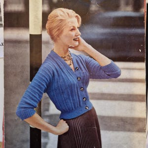 May include: Vintage knitting pattern featuring a woman wearing a blue cardigan and a brown pleated skirt. The pattern is for Motoravia Double-Knitting Wool, with bust sizes ranging from 32 to 42 inches. The Lee Target label is visible.