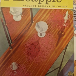 Puede incluir: Un libro de patrones de ganchillo vintage titulado "Pineapple Crochet Designs in Colour" de Coats. La portada presenta una foto de una mesa de madera con tres manteles individuales de ganchillo y un jarrón de vidrio con una vela.
