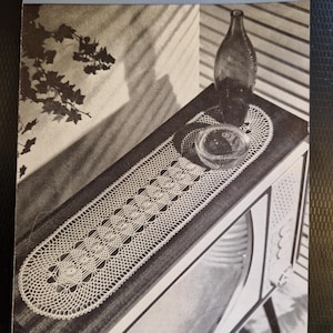 May include: A white crocheted table runner with a delicate floral pattern, resting on a wooden surface with a vintage television set in the background. The image is from a vintage crochet pattern booklet.