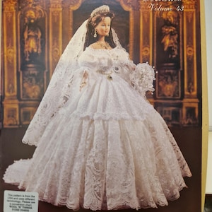 Può includere: Una bambola in un abito da sposa bianco all'uncinetto, velo e tiara, con un bouquet. L'immagine è tratta da "Crochet Collector Costume Volume 43" e presenta il testo "1840 Queen Victoria's Wedding Gown". Il modello include abito, strascico, bloomers, bouquet, scarpe e tiara.