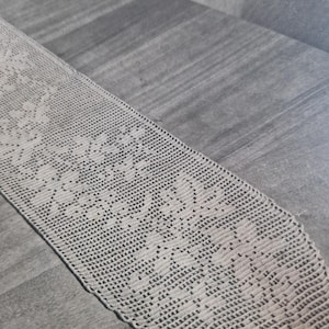 May include: A long, narrow, off-white crochet table runner featuring a repeating floral pattern created with intricate needlework. The textile has a delicate, open-weave texture and a scalloped edge design that runs along the length of the piece.