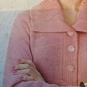 Puede incluir: Un cárdigan de punto rosa claro con cuello y tres botones grandes y redondos. El cuello presenta un diseño calado decorativo. Las mangas son largas y los brazos de la persona están cruzados.