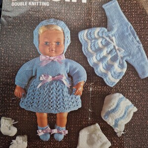 May include: A vintage knitting pattern booklet titled Robin 2631 Double Knitting featuring a doll wearing a light blue knitted dress, bonnet, and booties with pink ribbons, alongside a separate blue and white sweater, bonnet, and white pants.