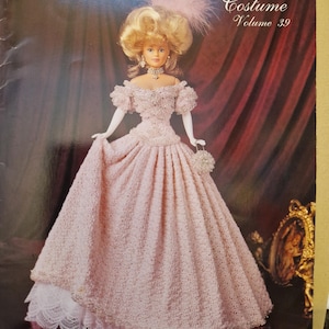 May include: A doll in a pink crochet gown with a matching purse and a blonde updo, featured on the cover of "Crochet Collector Costume Volume 39." The cover also includes the text "1896 Court Presentation Gown."