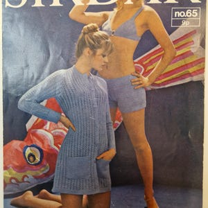 May include: Vintage magazine cover featuring two women modeling knitwear. One woman kneels in a light blue knit dress with pockets, while the other wears a matching blue knit bra and shorts. The magazine title "SIRDAR" is at the top.