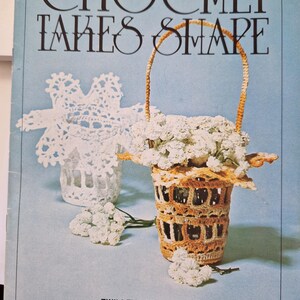 May include: A vintage crochet book titled "Crochet Takes Shape" with a photo of two crocheted baskets, one with a white handle and the other with a brown handle. Both baskets are filled with white flowers.