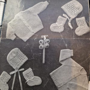 Puede incluir: Un folleto vintage para bebés titulado "BABY BOOKLET" de Hartfield, con ropa de bebé de punto. La imagen muestra una rebeca, un gorro, patucos y un sonajero. La portada del folleto es gris con texto e ilustraciones en negro.