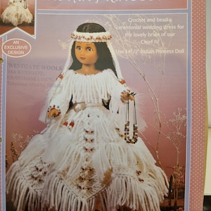 May include: A doll wearing a white crochet ceremonial wedding dress, with the text "INDIAN PRINCESS IV". The dress is adorned with fringe and bead accents. The doll is designed for a 14 1/2 inch doll.