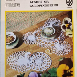 Może przedstawiać: Biała koronka szydełkowa z motywem łabędzia. Koronka leży na drewnianym stole z małą białą ceramiczną miską i czajnikiem. Obraz zawiera również kompozycję kwiatową z żółtymi i fioletowymi kwiatami.