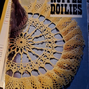 Op de afbeelding: Een vintage boekomslag met een gehaakt doily met een gerimpelde rand. De titel van het boek is "Ruffle Doilies" en de omslag is geel en bruin.