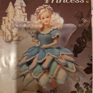 以下が含まれることがあります： 妖精のプリンセス人形の編み物パターン。人形は、花冠をつけた青、ピンク、白のドレスを着ています。パターンは「フェアリープリンセス」と呼ばれ、「GCP 21 To Knit」シリーズの一部です。