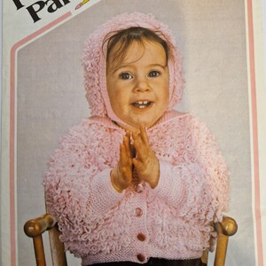 May include: A vintage knitting pattern cover featuring a baby wearing a pink hooded sweater. The sweater has a textured, looped design and button closure. The text "Peter Pan" and "Darling D.K." are visible. The pattern is for sizes 20-24 inches.