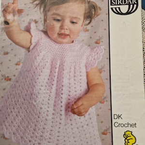 May include: A light pink crochet baby dress with a scalloped hem and cap sleeves. The dress is worn by a young child with pigtails. The image includes the text "DK Crochet" and the logo for Snuggly Baby Yarns.