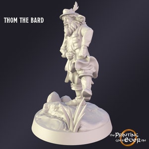 May include: A 3D-printed figurine of Thom the Bard, a character with a hat, feather, and period clothing. The figure stands on a small, rocky base with grass. The text "THOM THE BARD" is at the top left.