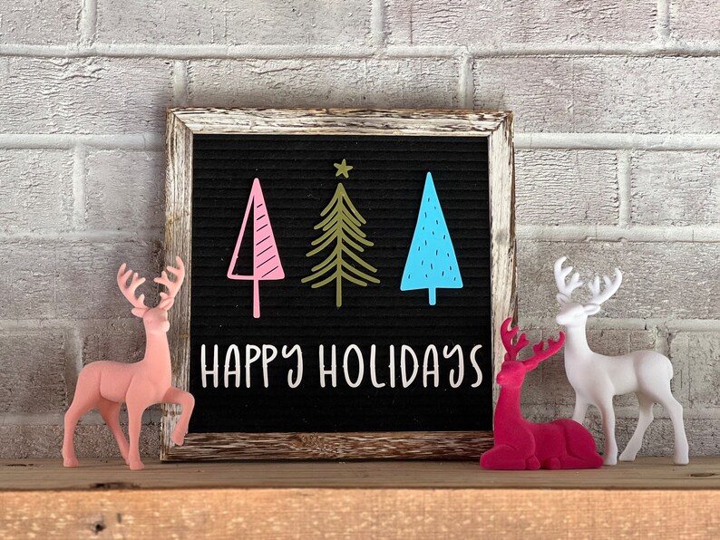 May include: A black letter board with a white wooden frame displays the message "Happy Holidays" with three stylized Christmas trees in pink, green, and blue. Two felt reindeer figurines, one pink and one white, are placed in front of the board.