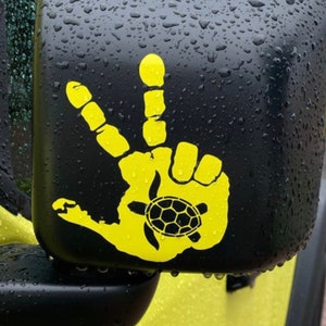 May include: A bright yellow vinyl decal applied to a black vehicle side mirror. The decal features a handprint shape forming a peace sign gesture, with a small black sea turtle silhouette centred within the palm area of the handprint design.