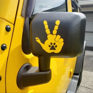 May include: A yellow vinyl decal applied to a black vehicle side mirror. The decal features a handprint design with two fingers raised in a peace sign, and a small black paw print centered within the palm area of the handprint graphic.