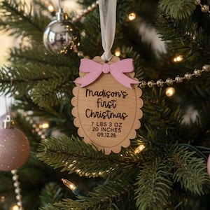 May include: A scalloped wooden ornament hanging from a white ribbon, featuring a light pink bow at the top. The text reads Madison's first Christmas, 7 LBS 3 OZ, 20 INCHES, and 09.12.26, engraved in black on the natural wood surface.