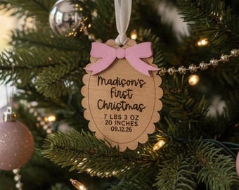 May include: A scalloped wooden ornament hanging from a white ribbon, featuring a light pink bow at the top. The text reads Madison's first Christmas, 7 LBS 3 OZ, 20 INCHES, and 09.12.26, engraved in black on the natural wood surface.