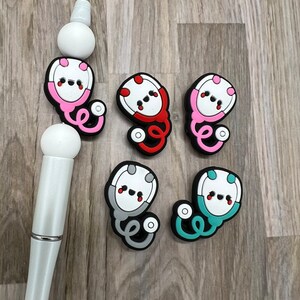 May include: Five colorful cartoon stethoscope charms with a white face and red cheeks. The charms are made of silicone and have a black outline. The charms are in pink, red, gray, teal, and blue.