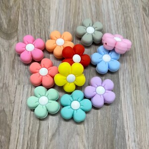 May include: Assortment of ten silicone flower-shaped beads in various colors, including pink, red, yellow, green, blue, purple, and gray. Each bead has a white center.