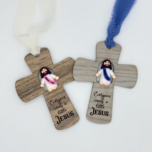 May include: Two wooden cross ornaments with a small Jesus figure and the text "Everyone needs a little Jesus." One cross has a white ribbon, the other a blue ribbon. The crosses have a wood grain pattern.