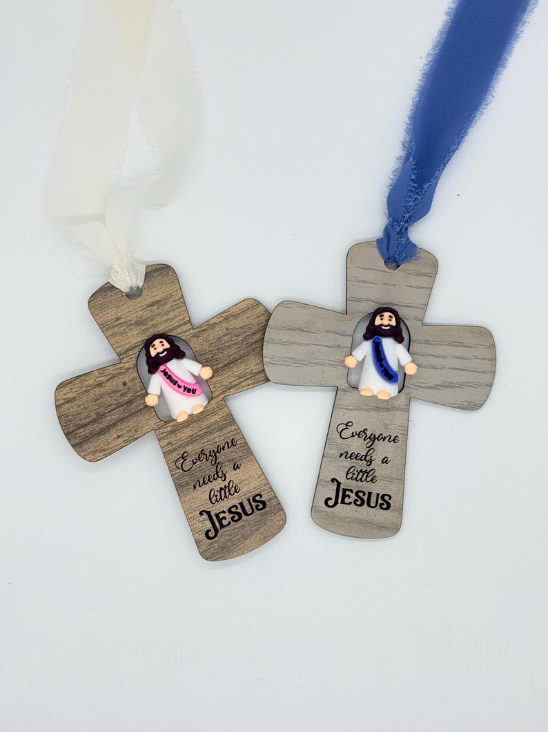 May include: Two wooden cross ornaments with a small Jesus figure and the text "Everyone needs a little Jesus." One cross has a white ribbon, the other a blue ribbon. The crosses have a wood grain pattern.
