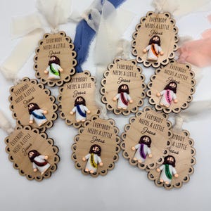 May include: Wooden ornaments with scalloped edges, each featuring a small Jesus figure with a colorful sash. The text "EVERYBODY NEEDS A LITTLE Jesus" is printed on each ornament. Ribbons in various colors are attached for hanging.