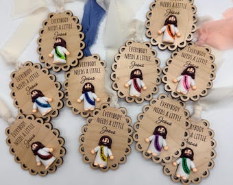 May include: Wooden ornaments with scalloped edges, each featuring a small Jesus figure with a colorful sash. The text "EVERYBODY NEEDS A LITTLE Jesus" is printed on each ornament. Ribbons in various colors are attached for hanging.
