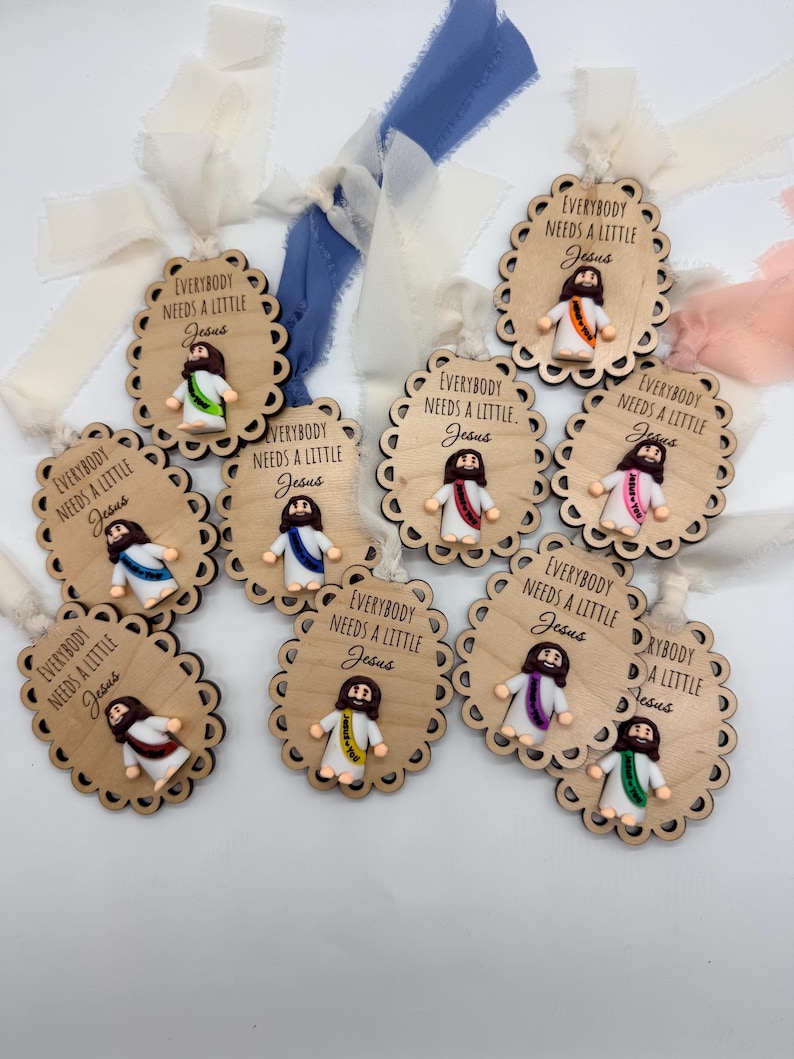May include: Wooden ornaments with scalloped edges, each featuring a small Jesus figure with a colorful sash. The text "EVERYBODY NEEDS A LITTLE Jesus" is printed on each ornament. Ribbons in various colors are attached for hanging.