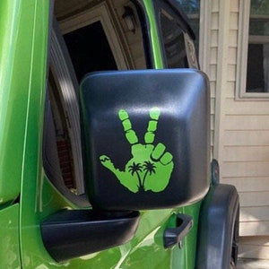 May include: A green Jeep Wrangler with a black side mirror. The mirror has a green decal of a hand with palm trees on it.